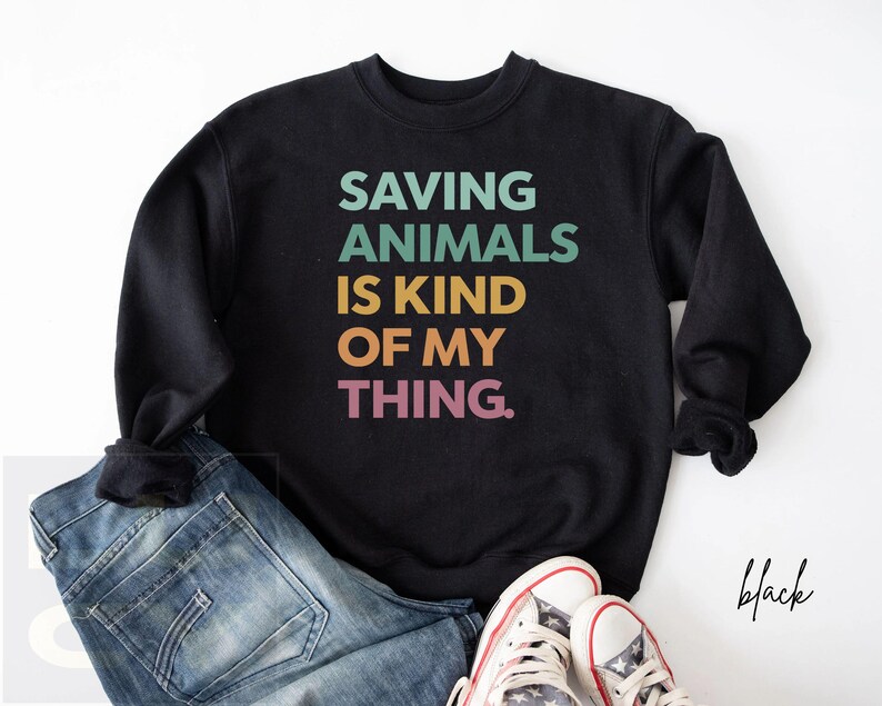 May include: Black sweatshirt with the text "SAVING ANIMALS IS KIND OF MY THING" in teal, green, yellow, and pink. The sweatshirt is paired with blue jeans and white sneakers.