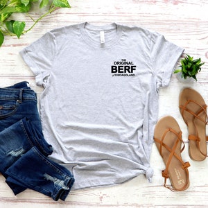 May include: A gray t-shirt with the text "THE ORIGINAL BERF of Chicagoland" printed on the front. The shirt is laid out on a white wooden surface with a pair of blue jeans and brown sandals.
