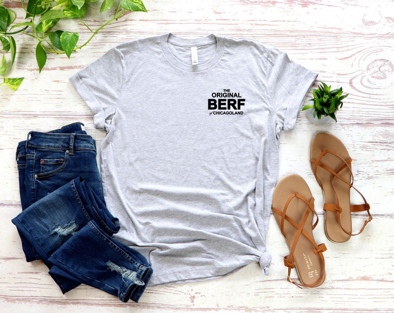 May include: A gray t-shirt with the text "THE ORIGINAL BERF of Chicagoland" printed on the front. The shirt is laid out on a white wooden surface with a pair of blue jeans and brown sandals.