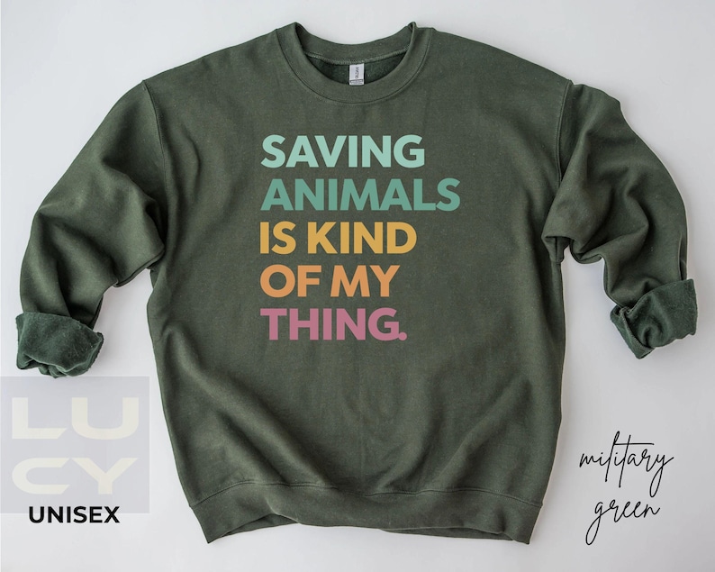 May include: A military green unisex sweatshirt with the text "SAVING ANIMALS IS KIND OF MY THING" in teal, light green, yellow, orange, and pink. The sweatshirt has long sleeves and a crew neck.