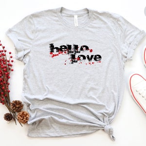 May include: A gray t-shirt with the text "hello love" in black with red splatters around the text. The shirt is on a white background with a sprig of red berries and pine cones.