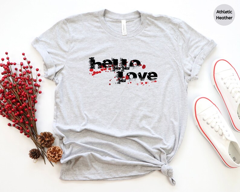 May include: A gray t-shirt with the text "hello love" in black with red splatters around the text. The shirt is on a white background with a sprig of red berries and pine cones.