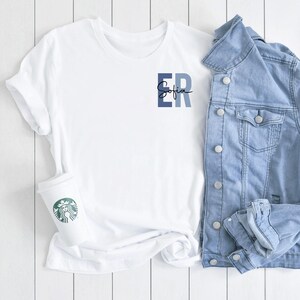 May include: White t-shirt with a blue and white "ER" monogram and the name "Sofia" written in cursive underneath. The t-shirt is paired with a light blue denim jacket.