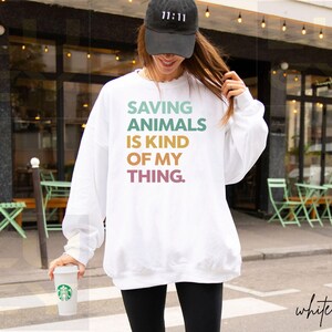May include: White sweatshirt with the text "SAVING ANIMALS IS KIND OF MY THING" in pastel colors. The person is wearing a black baseball cap with "11:11" on it and black leggings. A white coffee cup is held in the hand.