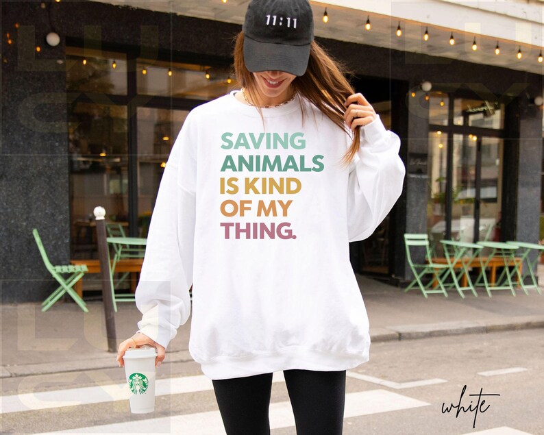 May include: White sweatshirt with the text "SAVING ANIMALS IS KIND OF MY THING" in pastel colors. The person is wearing a black baseball cap with "11:11" on it and black leggings. A white coffee cup is held in the hand.