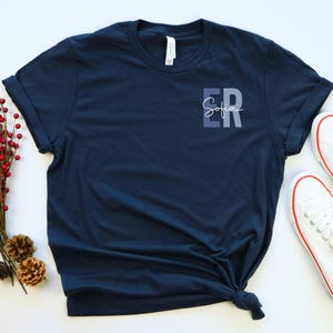 May include: A navy blue t-shirt with a pocket featuring the text "Sofia ER" in a gradient of blue shades.