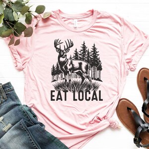 May include: Pink t-shirt with a black and white illustration of a deer in a forest with the text "Eat Local".