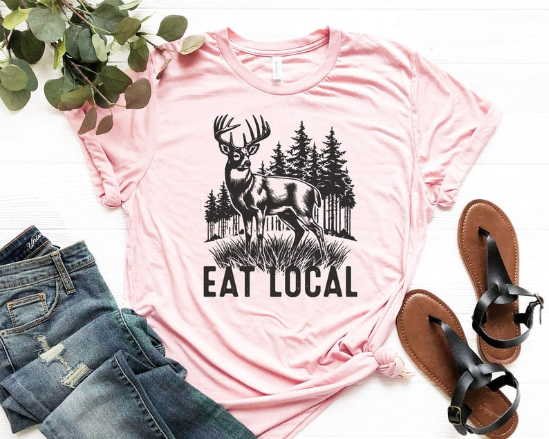 May include: Pink t-shirt with a black and white illustration of a deer in a forest with the text "Eat Local".