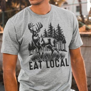 May include: A gray t-shirt with a black and white illustration of a deer in a forest setting. The text "Eat Local" is printed below the image.