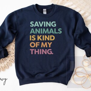 May include: Navy blue sweatshirt with the text "SAVING ANIMALS IS KIND OF MY THING" in teal, green, gold, orange, and pink. The sweatshirt has a crew neck and long sleeves. The word "navy" is written in cursive at the bottom left.