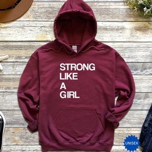 May include: Burgundy unisex hoodie with the text "STRONG LIKE A GIRL" in white lettering.