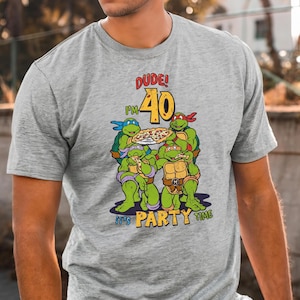 May include: A gray t-shirt featuring a graphic of the Teenage Mutant Ninja Turtles celebrating a 40th birthday with pizza. The text reads "DUDE! I'M 40" and "It's PARTY TIME" in yellow and red.