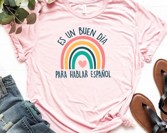 Op de afbeelding: Lichtroze t-shirt met de tekst "Es un buen día para hablar español" in turquoise en een regenboogafbeelding. De regenboog heeft rode, oranje, gele, turquoise en donker turquoise bogen met een roze hart in het midden.