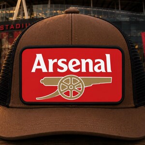 May include: A brown and black trucker hat featuring a rectangular patch on the front. The patch is red with a white Arsenal logo and a gold cannon graphic. The hat has a solid brown front panel and a black mesh back panel with a curved brim.