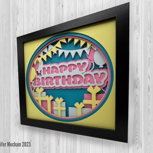 May include: A framed paper craft with a yellow background and a blue circle in the center. The circle features a pink and white "Happy Birthday" message with cut-out details of balloons, presents, and party flags.