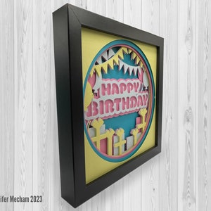 May include: A framed paper craft with a yellow background and a circle in the center. The circle has a blue background with a pink and white "Happy Birthday" banner. The banner is surrounded by white paper cutouts of presents and balloons.