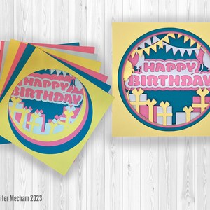 May include: A layered paper craft card with a circle design featuring the words "Happy Birthday" in pink letters with white outlines. The circle is surrounded by white and pink gift boxes, white balloons, and a white banner with flags.