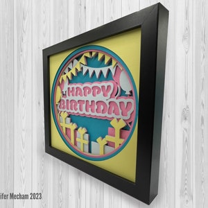 May include: A framed paper craft with a yellow background and a blue circle in the center. The circle has a pink and white "Happy Birthday" message with white and blue decorations around it.