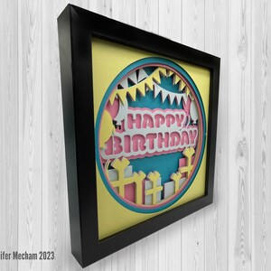 May include: A framed paper craft with a yellow background and a circle in the center. The circle has a blue background with a pink and white "Happy Birthday" message. The circle is surrounded by white and pink paper cutouts of balloons, streamers, and presents.