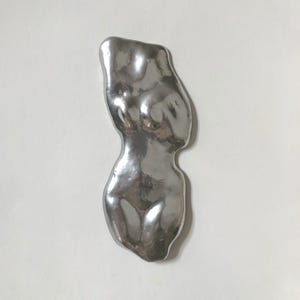 May include: A silver-colored, abstract sculpture of a human torso. The sculpture has a smooth, reflective surface and is set against a white background. The form is stylized, with rounded shapes suggesting the curves of the body.