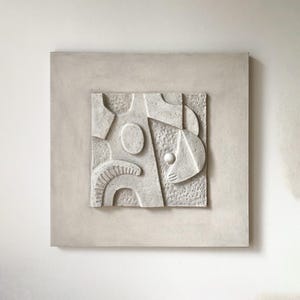 May include: An off-white relief sculpture with an abstract design, presented within a square frame. The artwork features a textured surface with geometric shapes, including ovals and curved lines. The frame and sculpture share a similar colour and texture.