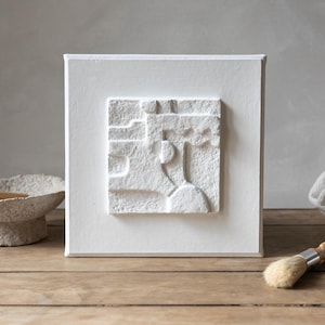 Peut inclure: Une œuvre d'art murale texturée blanche avec un motif géométrique en relief. L'œuvre est encadrée dans un cadre blanc et est exposée sur une surface en bois.