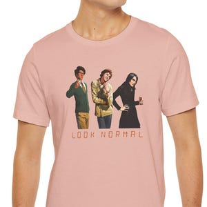 Peut inclure: T-shirt rose clair avec un motif de trois personnes en vêtements vintage. Le texte "LOOK NORMAL" est imprimé en dessous dans une police sans-serif. Le t-shirt est fait d'une matière douce.