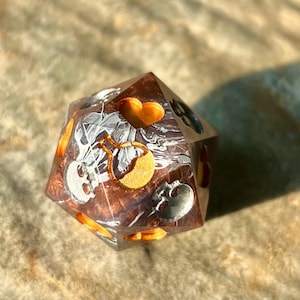 May include: A multi-sided, brown and clear resin die with embedded silver skulls, gold hearts, and a potion bottle design. The die has orange accents and a marbled interior, suitable for tabletop gaming.