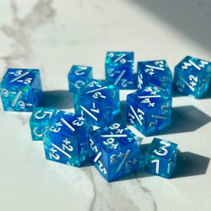 May include: A collection of twelve blue and clear resin dice with white numbering and symbols. The dice are scattered across a light-colored surface, with some faces showing fractional numbers and plus/minus symbols.