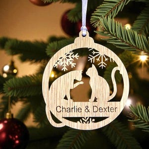 May include: Wooden Christmas bauble with a cat silhouette design and the names "Charlie & Dexter" engraved. The bauble is hanging on a Christmas tree with other decorations and lights.