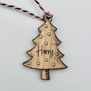 K&ouml;nnte beinhalten: Weihnachtsbaumschmuck aus Holz in Form eines Baumes, mit dem eingravierten Namen "Harry". Der Baum hat einen Stern an der Spitze und ist mit kleinen Kreisen verziert. Er h&auml;ngt an einer rot-wei&szlig;-gr&uuml;nen Schnur.