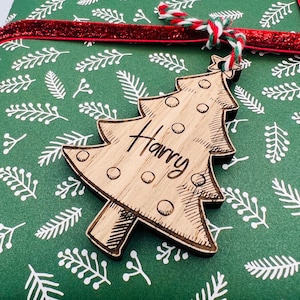 K&ouml;nnte beinhalten: Ein Weihnachtsbaum-Geschenkanh&auml;nger aus Holz mit dem eingravierten Namen "Harry". Der Anh&auml;nger ist an einem Geschenk befestigt, das in gr&uuml;nes Papier mit wei&szlig;em Bl&auml;ttermuster und einem roten Band verpackt ist. Der Anh&auml;nger ist aus hellem Holz.