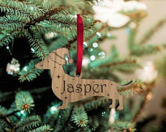 Puede incluir: Un adorno navideño de madera con forma de dachshund que lleva un gorro de Papá Noel. El adorno está personalizado con el nombre "Jasper".