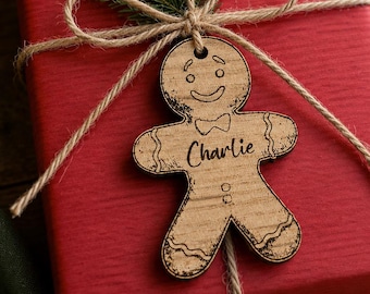 Könnte beinhalten: Ein rotes Geschenkpaket, das mit Schnur gebunden ist und mit einem hölzernen Lebkuchenmann-Anhänger mit dem Namen "Charlie" verziert ist. Die Box ist mit Tannenzweigen und roten Beeren dekoriert.