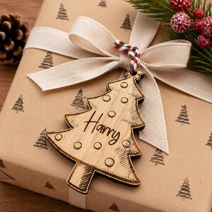 K&ouml;nnte beinhalten: Ein Weihnachtsgeschenk, verpackt mit braunem Papier und einem beigen Band, verziert mit einem h&ouml;lzernen Weihnachtsbaum-Anh&auml;nger. Der Anh&auml;nger ist mit dem Namen "Harry" graviert. Das Geschenkpapier hat ein sich wiederholendes Baummuster.