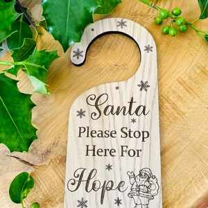 May include: Wooden door hanger with the text "Santa Please Stop Here For Hope" and snowflake accents. A small illustration of Santa Claus is at the bottom. The hanger is light brown and has a curved top for hanging.