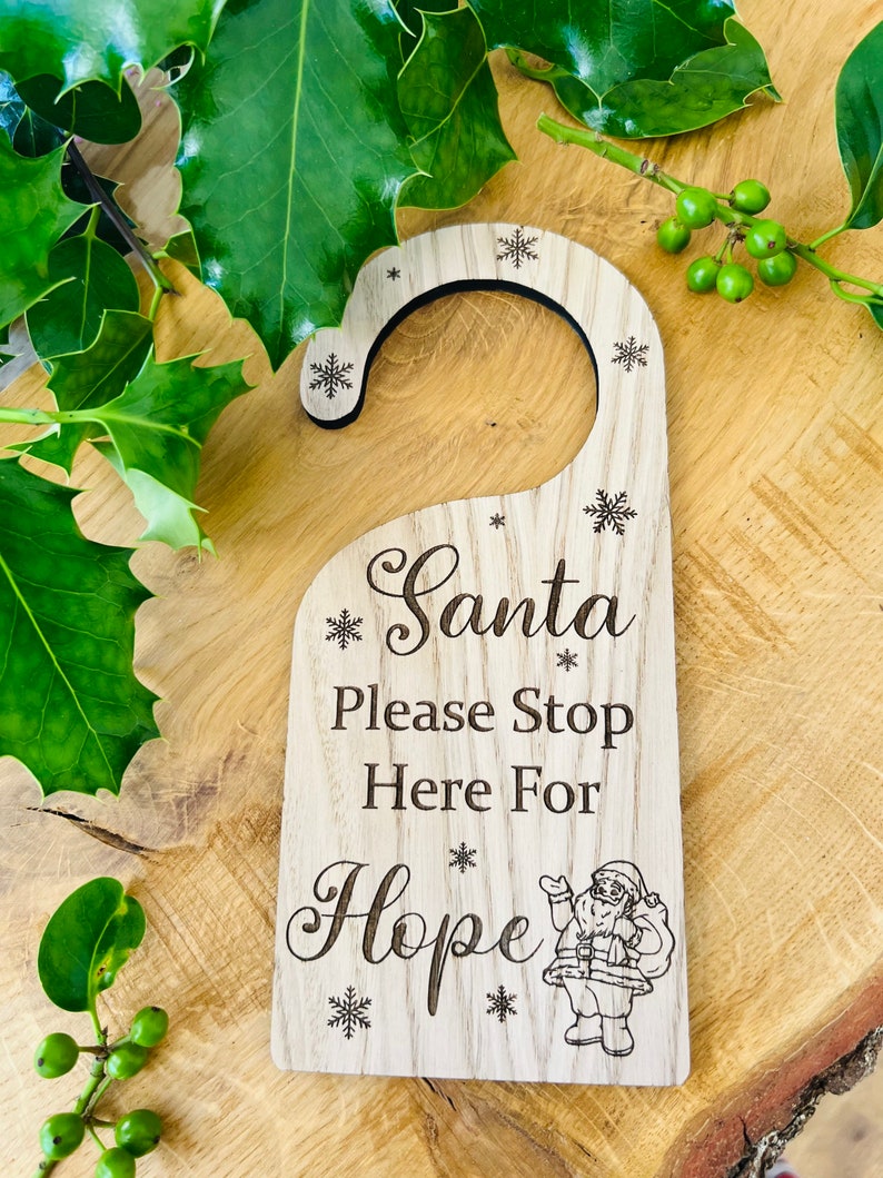 May include: Wooden door hanger with the text "Santa Please Stop Here For Hope" and snowflake accents. A small illustration of Santa Claus is at the bottom. The hanger is light brown and has a curved top for hanging.