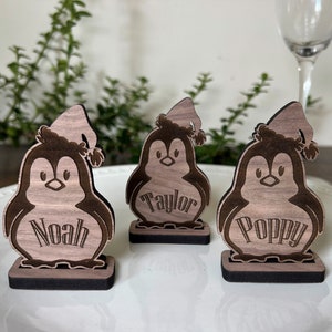 Könnte beinhalten: Drei Holzpinguin-Figuren mit den eingravierten Namen: Noah, Taylor und Poppy. Jeder Pinguin trägt eine Weihnachtsmütze. Die Pinguine stehen auf einem weißen Teller, mit Grünzeug im Hintergrund.