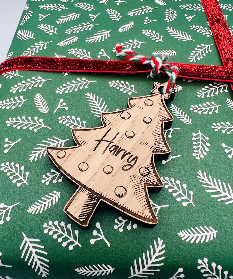 K&ouml;nnte beinhalten: Ein Weihnachtsbaum-Geschenkanh&auml;nger aus Holz mit dem eingravierten Namen "Harry". Der Anh&auml;nger ist an einem Geschenk befestigt, das in gr&uuml;nes Papier mit wei&szlig;em Bl&auml;ttermuster und einem roten Band verpackt ist. Der Anh&auml;nger ist aus hellem Holz.