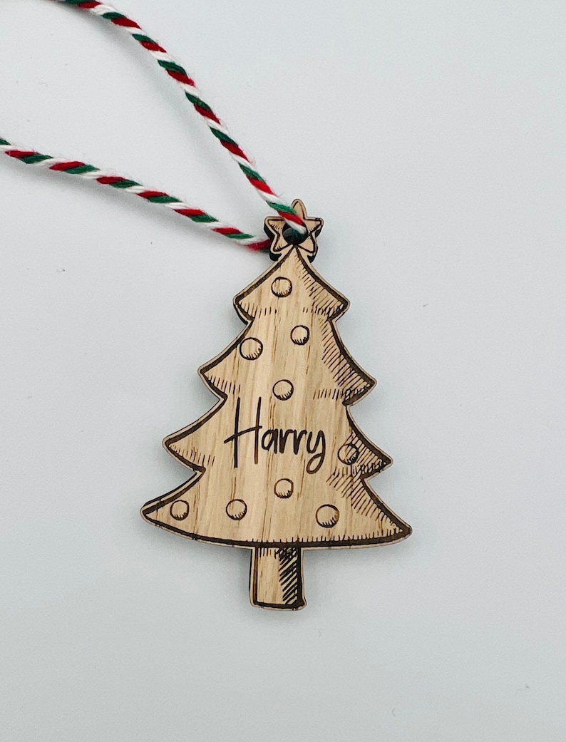 K&ouml;nnte beinhalten: Weihnachtsbaumschmuck aus Holz in Form eines Baumes, mit dem eingravierten Namen "Harry". Der Baum hat einen Stern an der Spitze und ist mit kleinen Kreisen verziert. Er h&auml;ngt an einer rot-wei&szlig;-gr&uuml;nen Schnur.