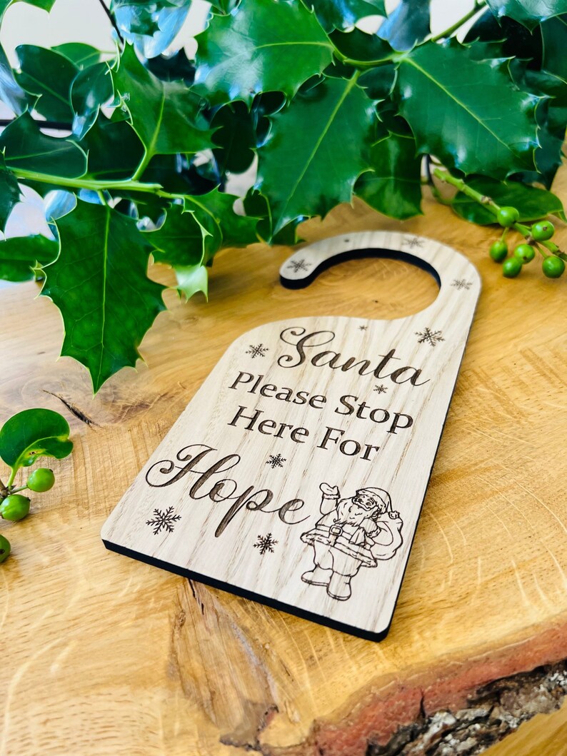 May include: A wooden door hanger with the text "Santa please stop here for Hope" and a Santa Claus illustration. The door hanger is light brown with a black border and snowflake details. The background includes green holly.