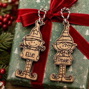 Può includere: Due decorazioni in legno a forma di elfo con dettagli incisi, ciascuna dotata di un cartello con i nomi Ellie e Freddie. Gli ornamenti sono appesi a cordini intrecciati rossi, bianchi e verdi e presentano una finitura in legno naturale.