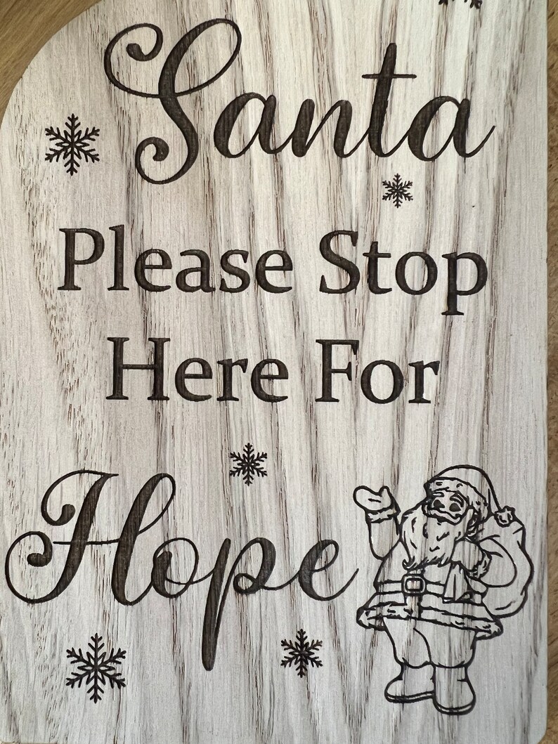 May include: Wooden sign with the words "Santa Please Stop Here For Hope" in a decorative font. The sign has a light wood grain background with engraved snowflakes and a Santa Claus illustration.