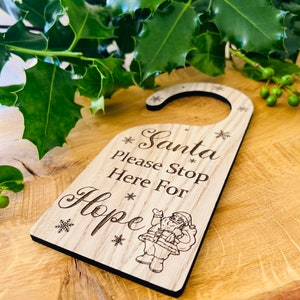 May include: Wooden door hanger with the text "Santa Please Stop Here For Hope" and a Santa Claus illustration. The hanger is light brown with a black border and snowflake accents, perfect for Christmas.