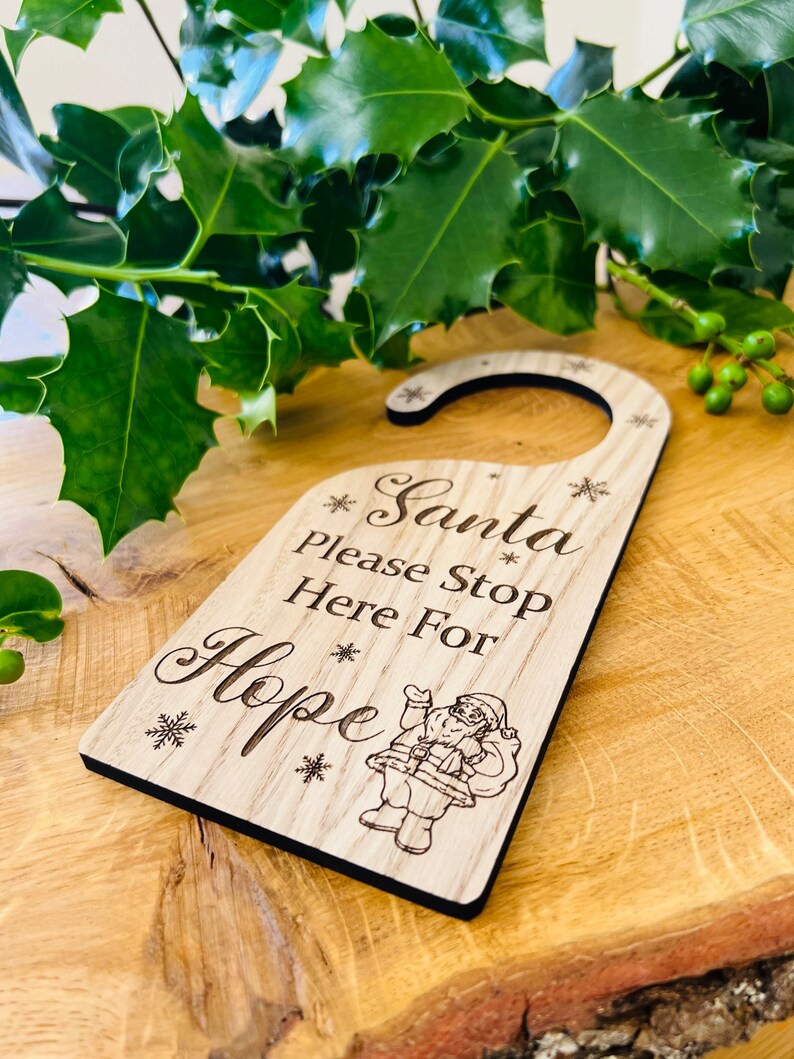 May include: Wooden door hanger with the text "Santa Please Stop Here For Hope" and a Santa Claus illustration. The hanger is light brown with a black border and snowflake accents, perfect for Christmas.