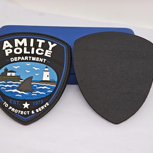May include: Two shield-shaped patches, one featuring a black and blue design with the text AMITY POLICE DEPARTMENT EST. 1975 TO PROTECT & SERVE, a lighthouse, a pier, and a shark fin in the ocean, and the other patch is a solid black shield shape.