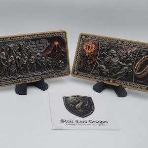 May include: Two rectangular metal plaques featuring detailed relief scenes from fantasy literature, displayed on black stands. One plaque shows a group of figures walking toward a volcano with the text "Not all those who wander are lost". The other shows a glowing eye and a ring. A card with a dragon shield logo and "Stoic Coin Designs" is included.