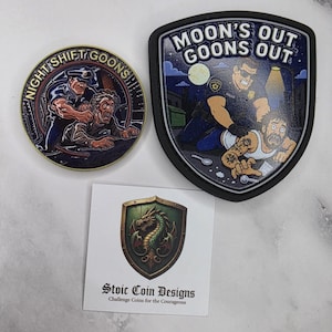 May include: A circular challenge coin with the text NIGHT SHIFT GOONS, a shield-shaped patch with the text MOON'S OUT GOONS OUT, and a square card featuring a dragon crest and the text Stoic Coin Designs Challenge Coins for the Courageous.