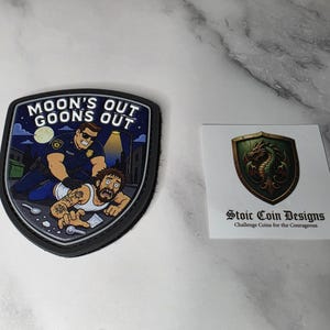 Op de afbeelding: Een schildvormige patch met een cartoon politieagent die een persoon arresteert met de tekst MOON'S OUT GOONS OUT, naast een vierkante witte kaart met een groen draak-schildlogo en de tekst Stoic Coin Designs.