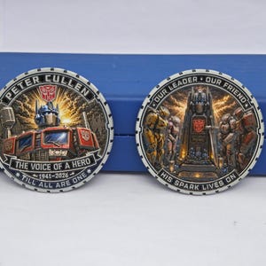 Peut inclure: Deux pièces commémoratives circulaires représentant Optimus Prime. La pièce de gauche porte les inscriptions PETER CULLEN, THE VOICE OF A HERO, 1941-2026, TILL ALL ARE ONE. Celle de droite affiche OUR LEADER, OUR FRIEND, HIS SPARK LIVES ON.